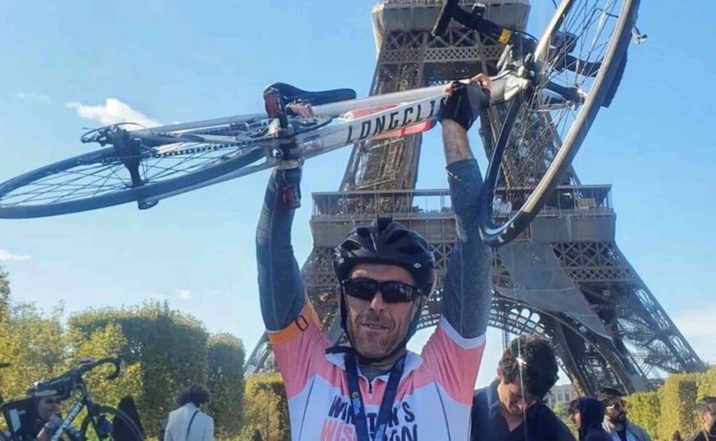 Cyclist in a Winston's Wish top holding his bike above his head in front of the Eiffel Tower