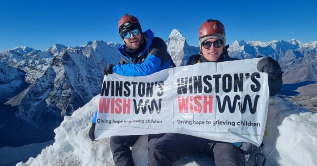 Two Winston's Wish supporters at the top of a snowy mountain with a Winston's Wish banner