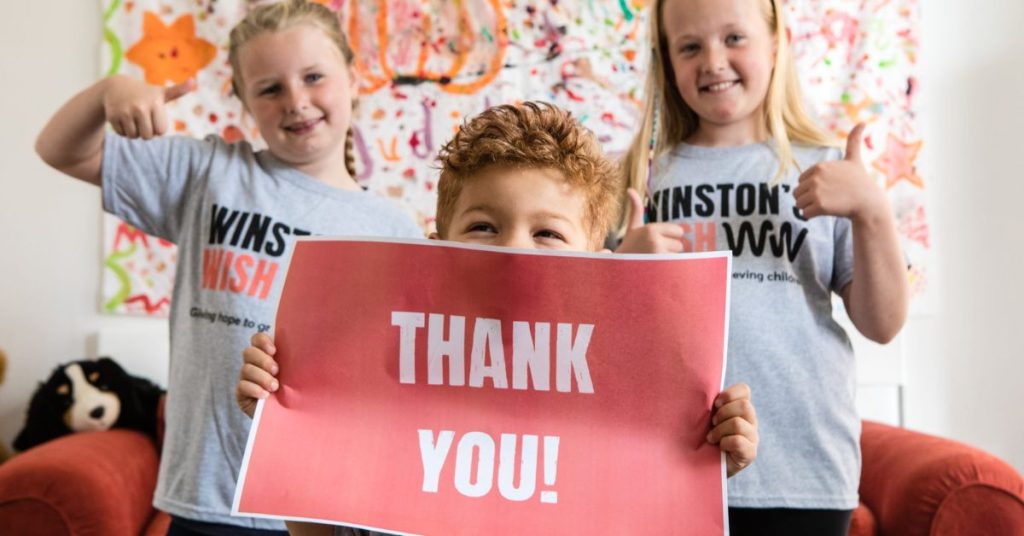 Three young children in Winston Wish t-shirts with a sign that reads 'thank you.'